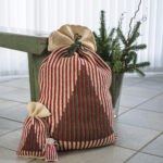 Hæklede julesække i tre størrelser med lodrette røde, grønne og cremefarvede striber. De to største julesække har et mønstret juletræ på forsiden, og alle tre er bundet foroven. Sækkene står på et gulv ved en grøn bænk og en vase med gran, som en del af en hyggelig juledekoration.