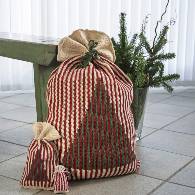Hæklede julesække i tre størrelser med lodrette røde, grønne og cremefarvede striber. De to største julesække har et mønstret juletræ på forsiden, og alle tre er bundet foroven. Sækkene står på et gulv ved en grøn bænk og en vase med gran, som en del af en hyggelig juledekoration.
