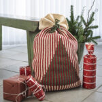 Hæklede julesække i tre størrelser med lodrette røde, grønne og cremefarvede striber. De to største julesække har et mønstret juletræ på forsiden, og alle tre er bundet foroven. Sækkene står på et gulv ved en grøn bænk og en vase med gran, som en del af en hyggelig juledekoration.