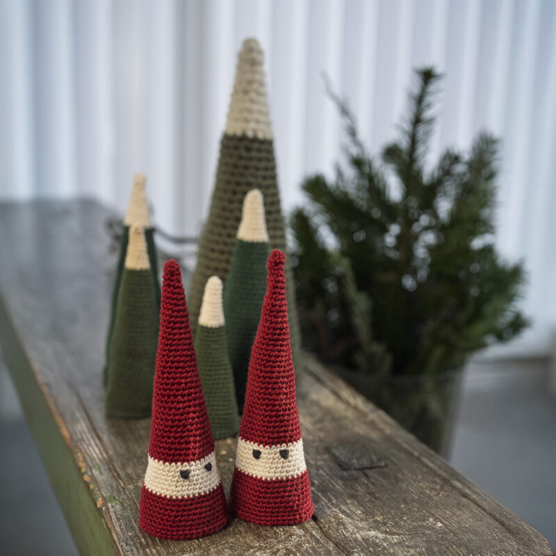 Hæklede keglenisser i rød og naturfarve og keglejuletræer i grønne nuancer står samlet på et træbord som minimalistisk julepynt.