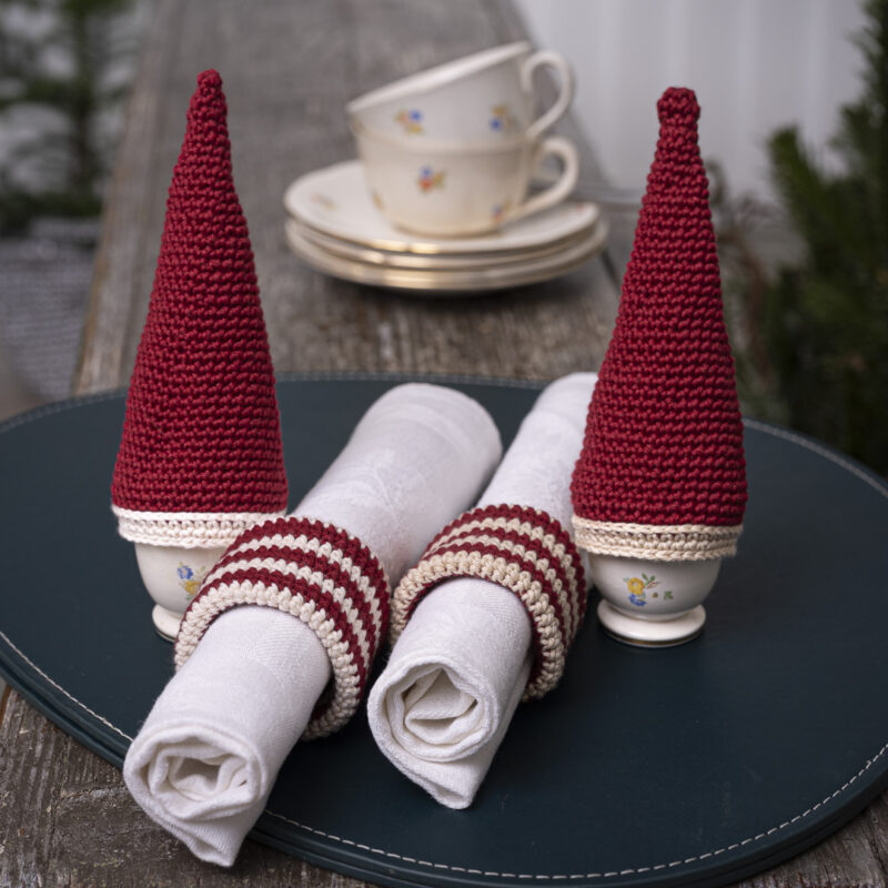 Håndhæklede kegleformede æggevarmere i rød med lys kant, placeret på æggebægre, sammen med matchende stribede servietringe i rød og creme omkring hvide stofservietter på et julebord.